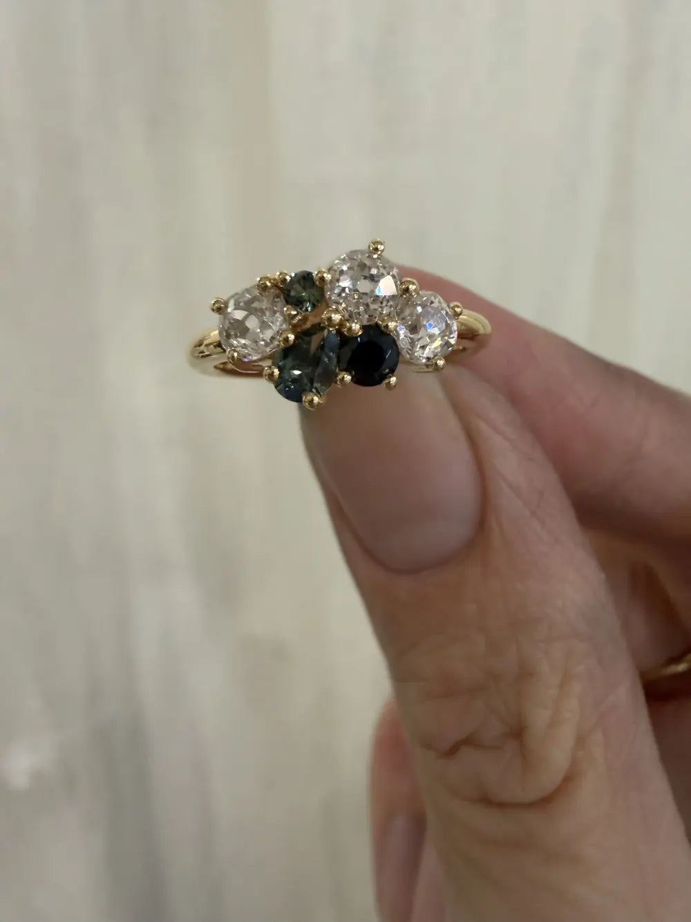 A bespoke cluster ring with old-cut diamonds and green and blue sapphires, held between finger and thumb