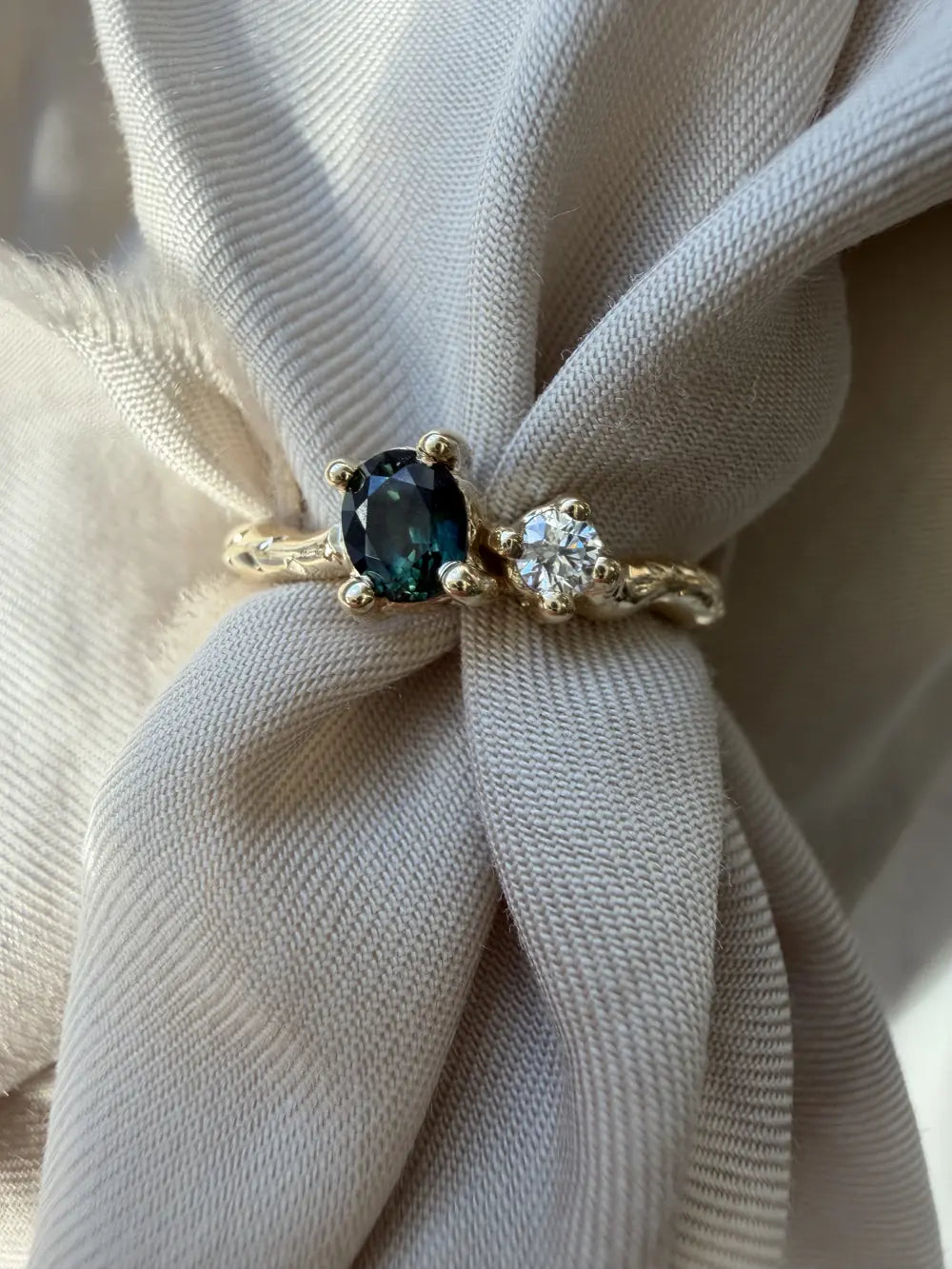 A bespoke ring with a teal sapphire and a round diamond, resting in grey fabric